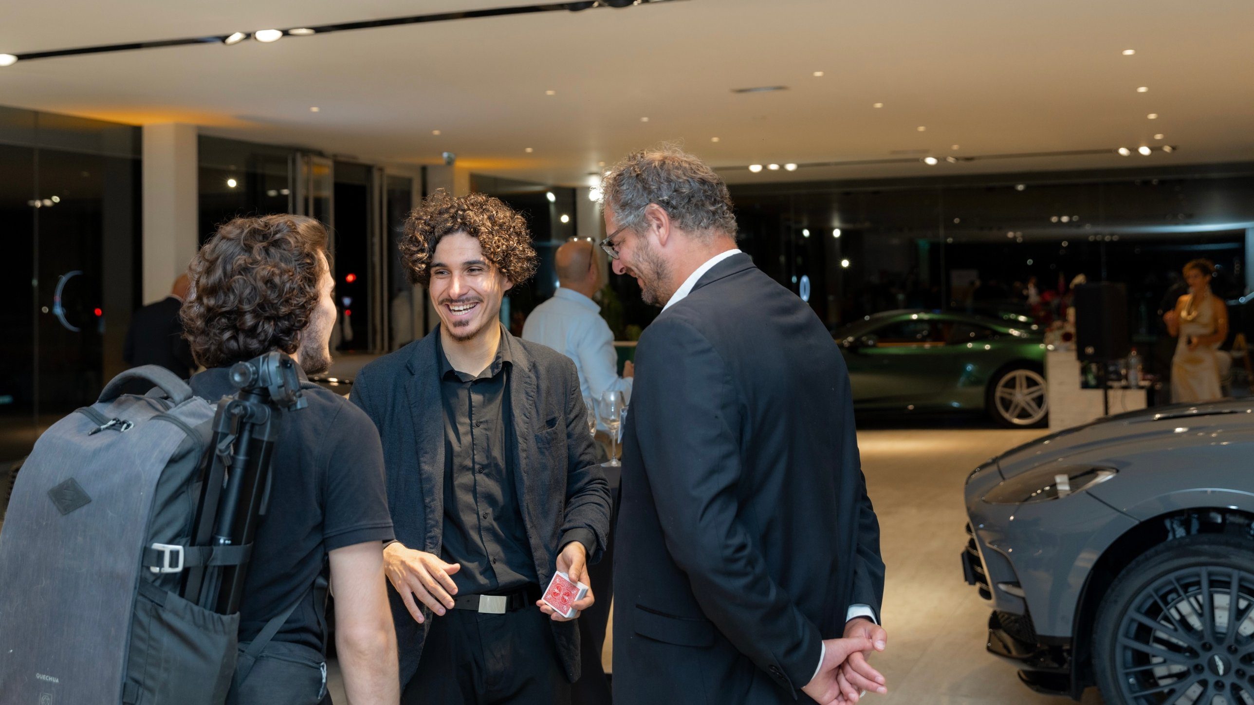 Mehdi Hideche, souriant, jeu de cartes en main, avec des invités dans une concession Aston Martin